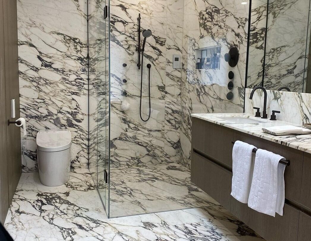 Modern bathroom with glass shower, white marble walls and floor, black shower fixtures, and a floating vanity with dual sinks.